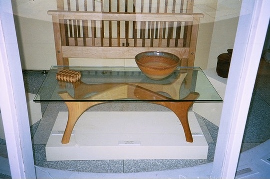 A glass-topped coffee table with sculpted legs in a display case, a slatted headboard behind it